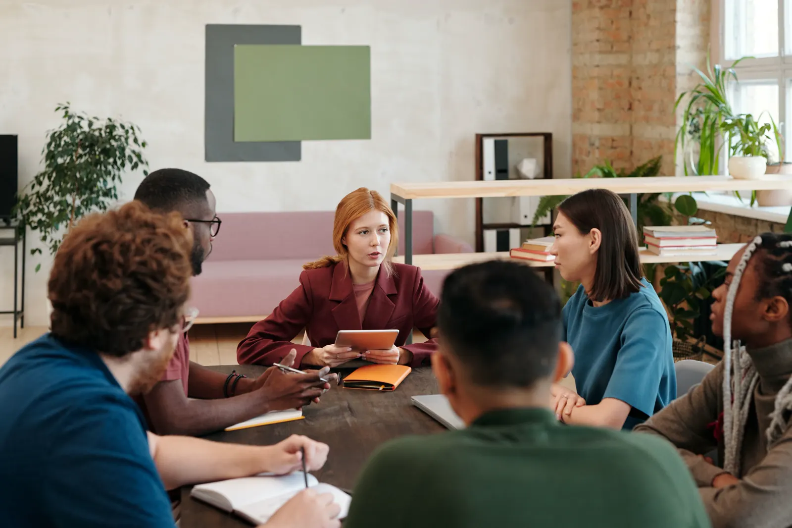 Six colleagues talking around a table in a bright, plant-filled workroom, one of them holding a tablet.